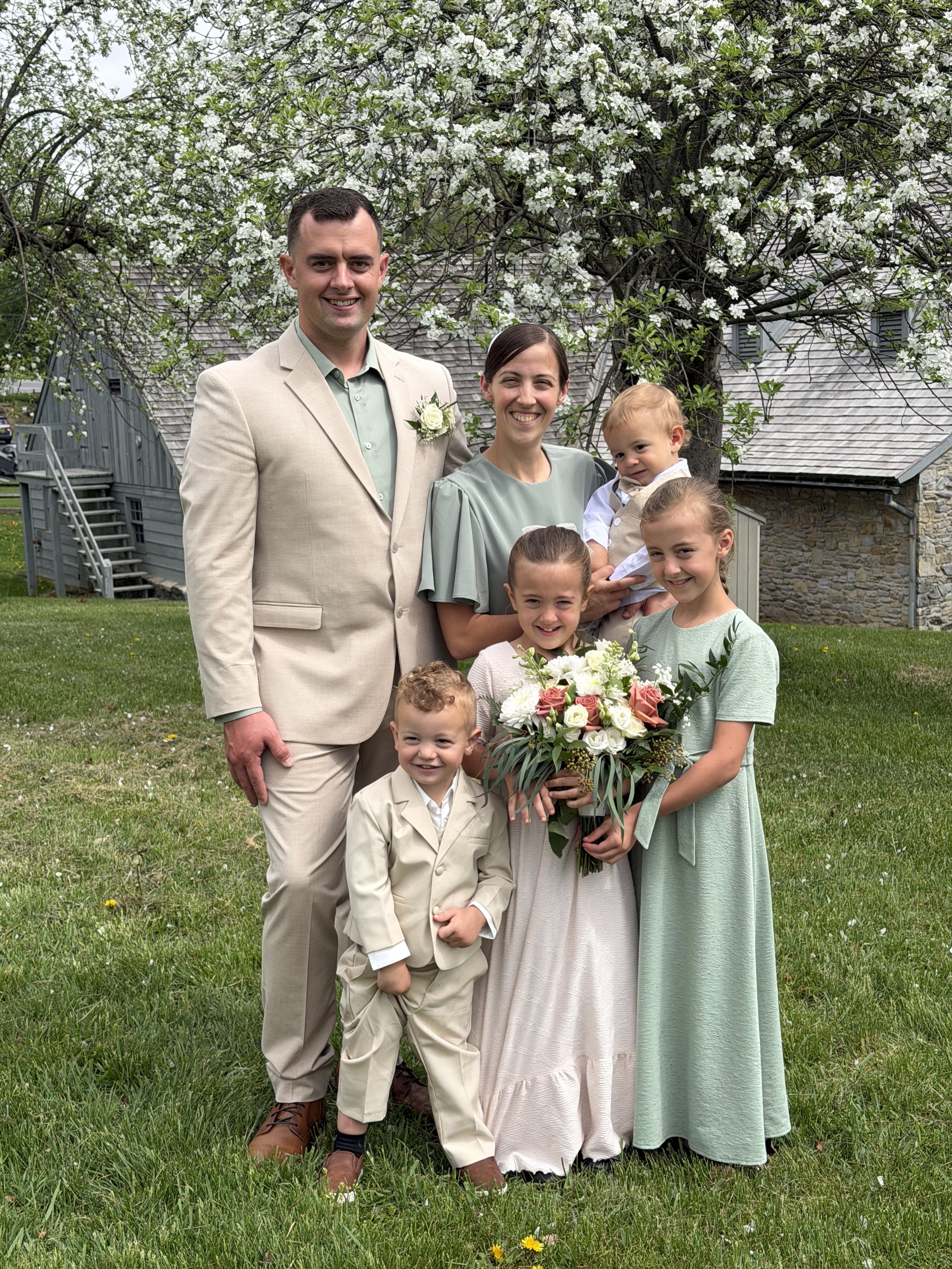 A family of seven standing on green grass in front of a flowering tree and a building, celebrating a special occasion, with some members holding flowers.
