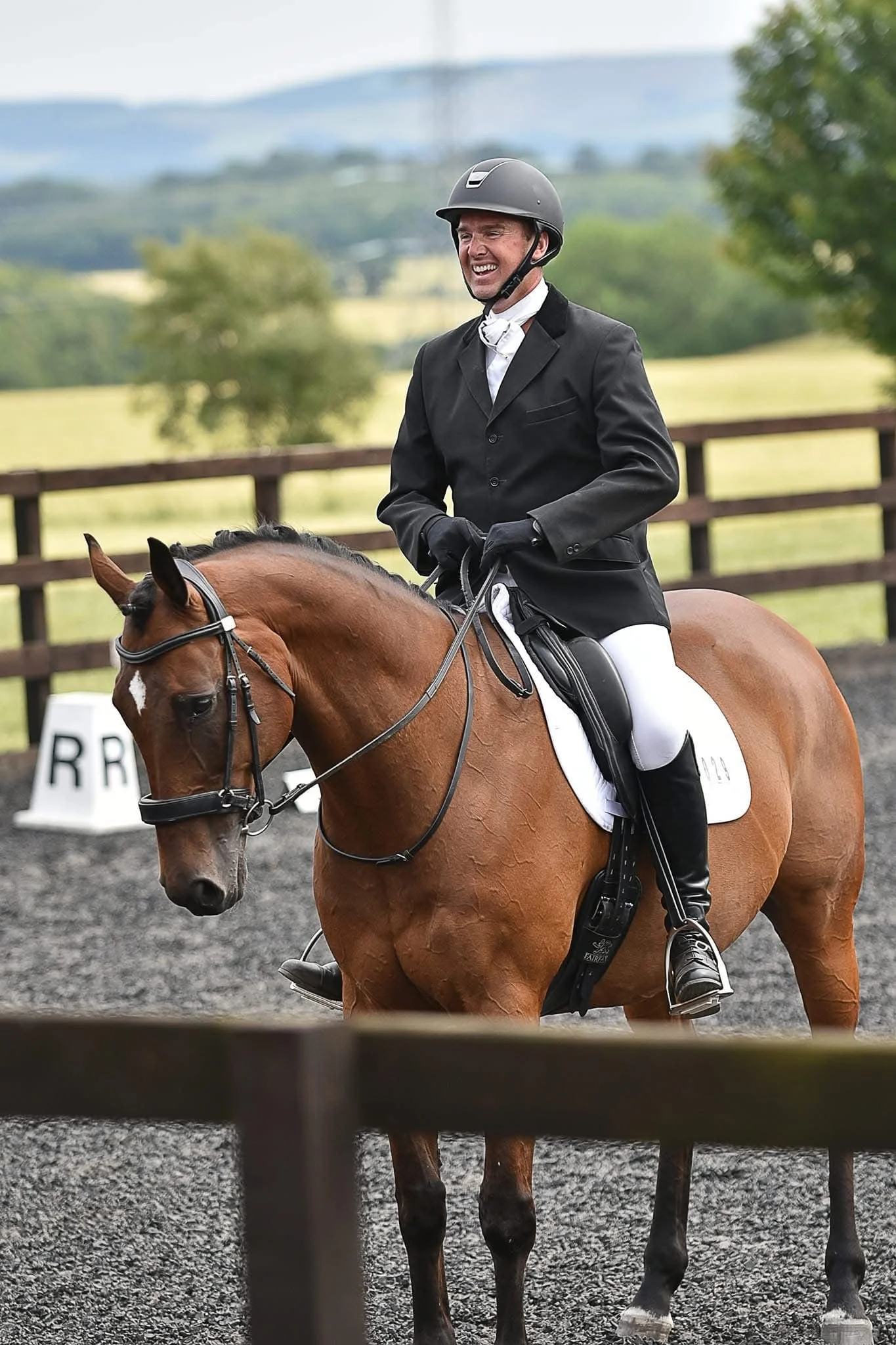 A smiling man in a riding helmet and black jacket standing next to a brown horse with a white star on its forehead, inside a stable.