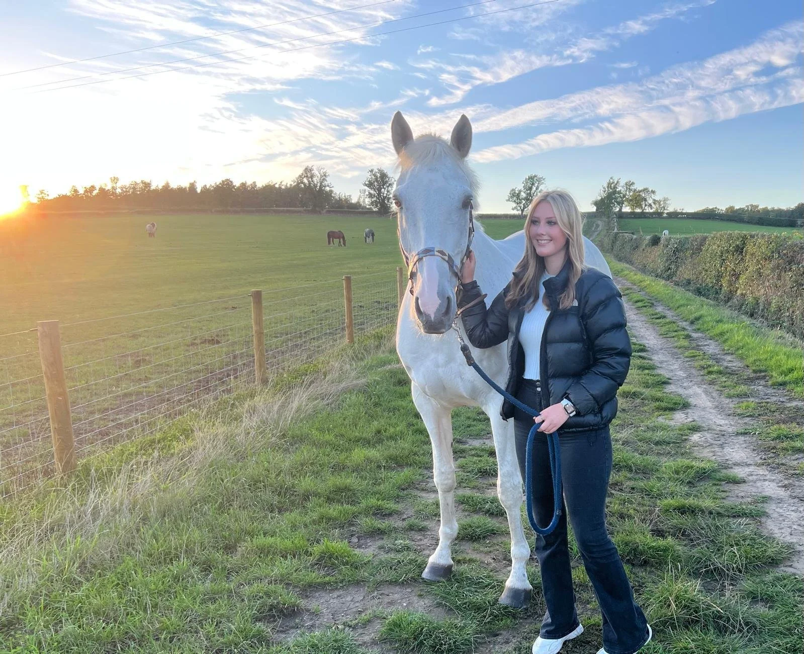 A young woman with long, blonde hair, wearing a black puffy jacket, white sweater, and dark jeans, holding a white horse by the bridle, standing on a dirt path in a green field during sunset.
