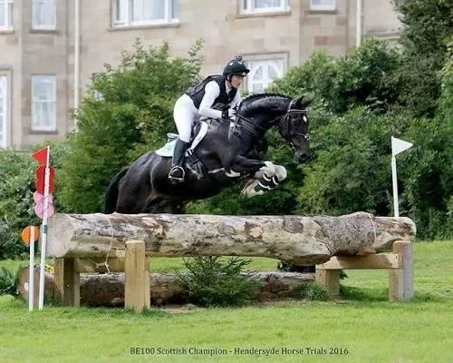 A woman smiling, taking a selfie with a white horse inside a stable.