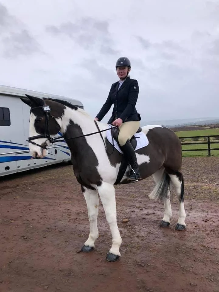 A woman dressed in equestrian riding attire, including a helmet, jacket, and riding boots, sitting on a black and white pinto horse in an outdoor riding arena with a cloudy sky and fencing in the background.