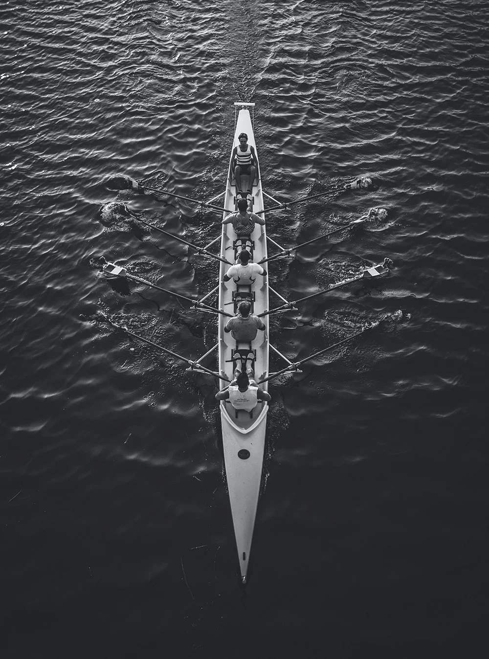 Top view of a rowing team in a boat on water, with four rowers using oars in unison, and a coxswain at the front.