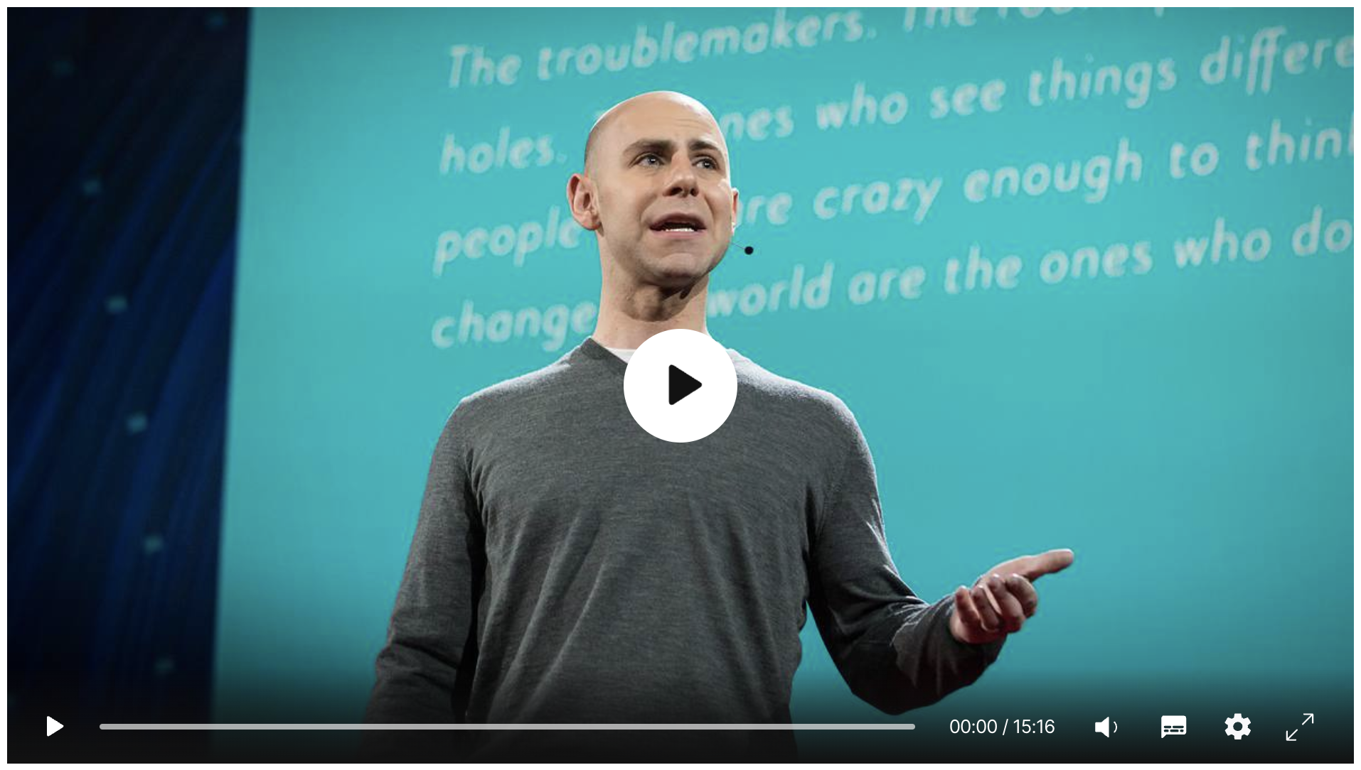 A person speaking on stage with a headset in front of a blue screen displaying text, during a presentation or TED Talk.