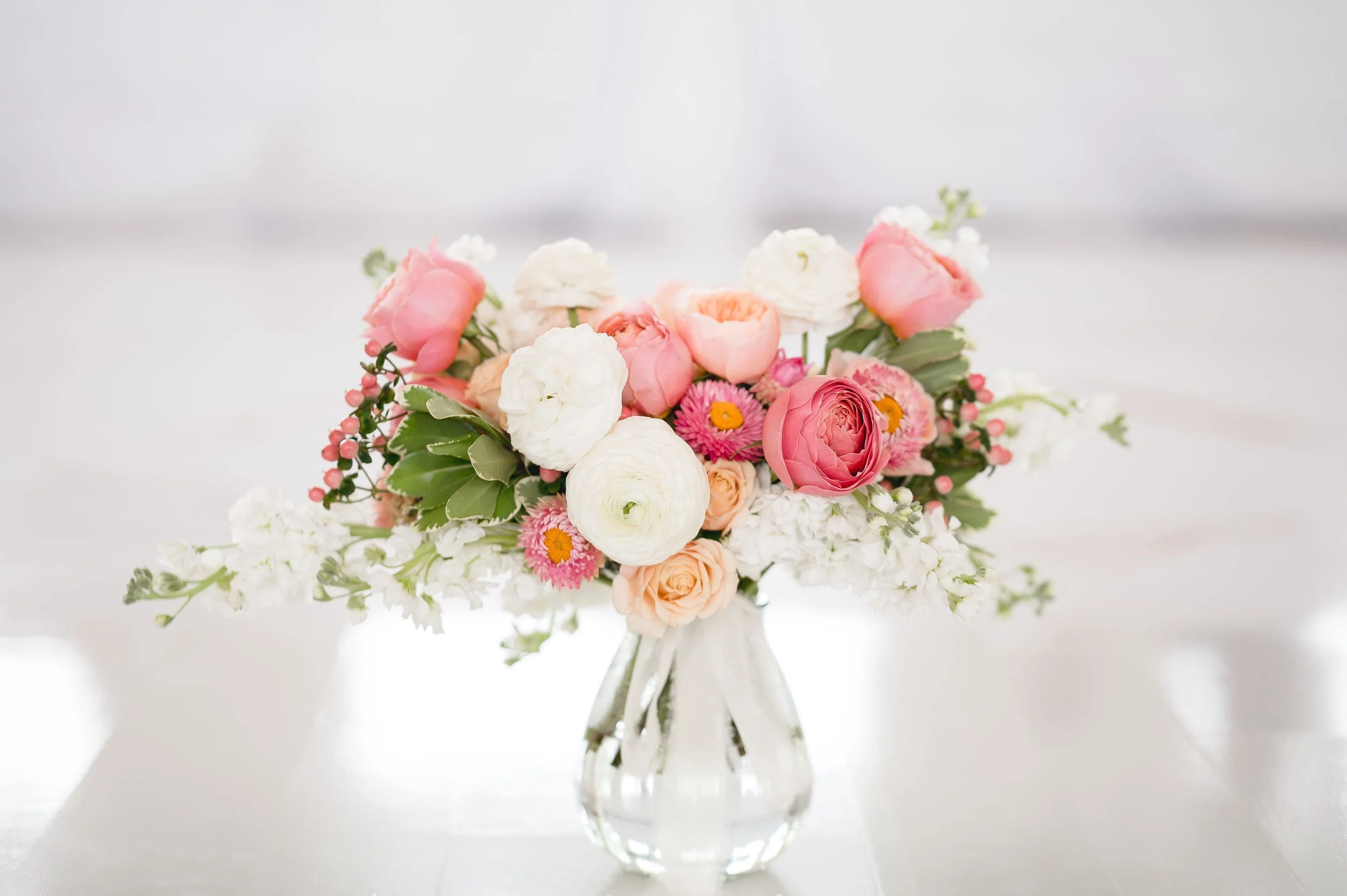 Pink, white, and peach floral bouquet in a clear glass vase on a white surface.