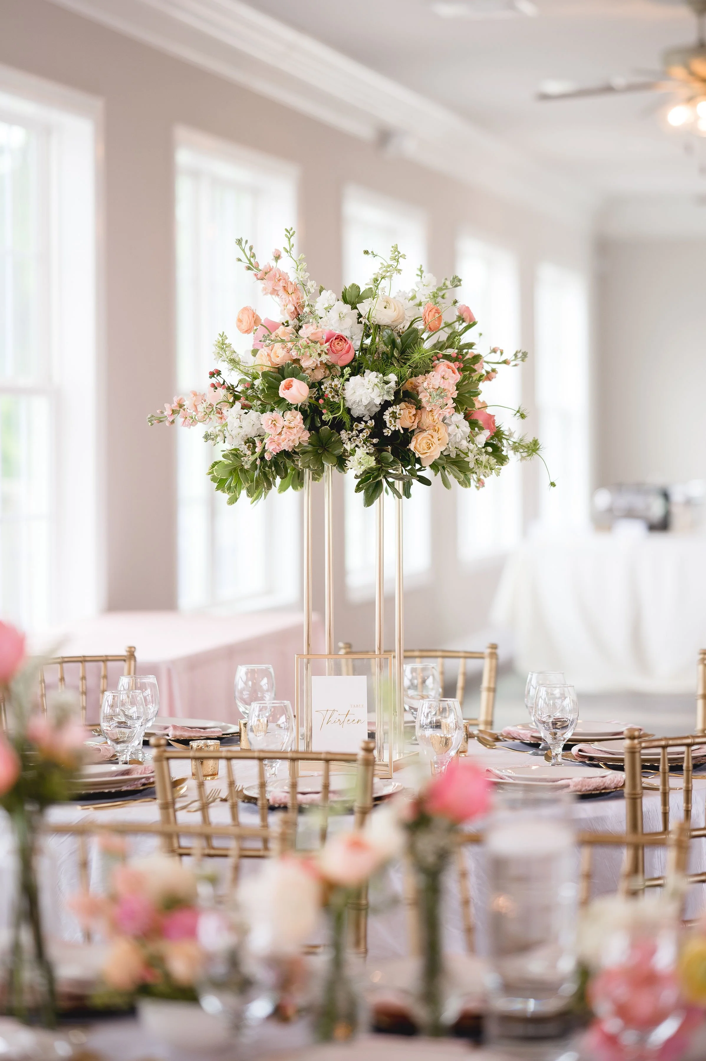 Elegant table centerpiece with a large floral arrangement of pink, white, and cream flowers on a gold stand at a formal event.