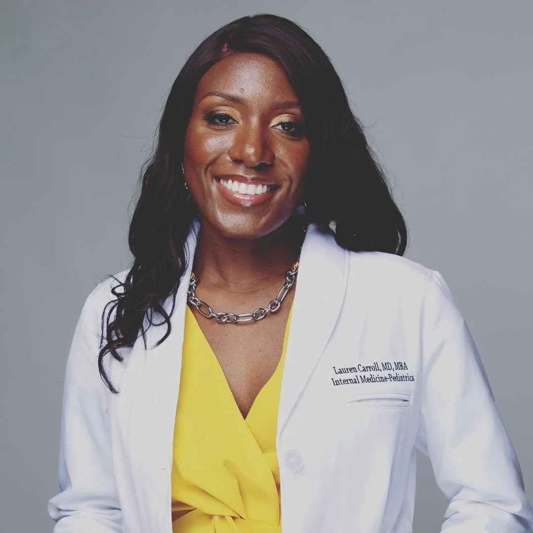 A woman with long black hair, smiling, wearing a white medical coat with name and title, yellow blouse, and silver chain necklace against a gray background.