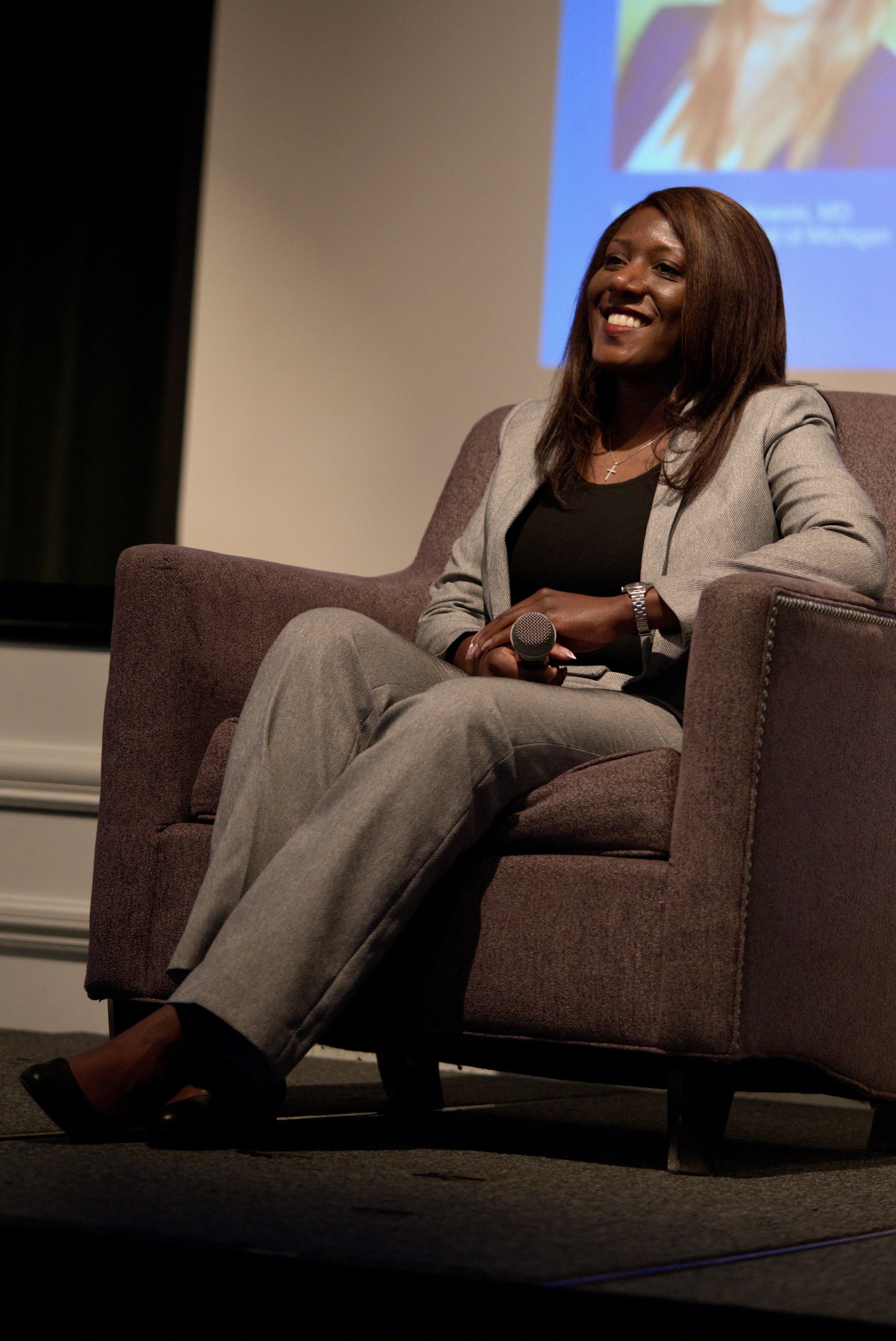 A woman in a grey suit sitting on a cushioned chair, holding a microphone, smiling during a presentation or panel discussion, with a projection screen in the background.