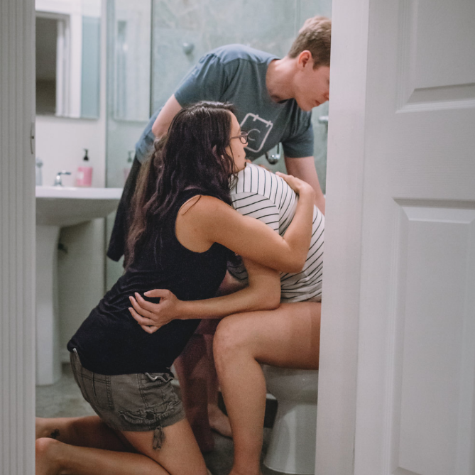 A birth doula and supportive partner comforting a laboring woman in a bathroom setting, representing hands-on, personalized birth support services offered by Baby Caravan