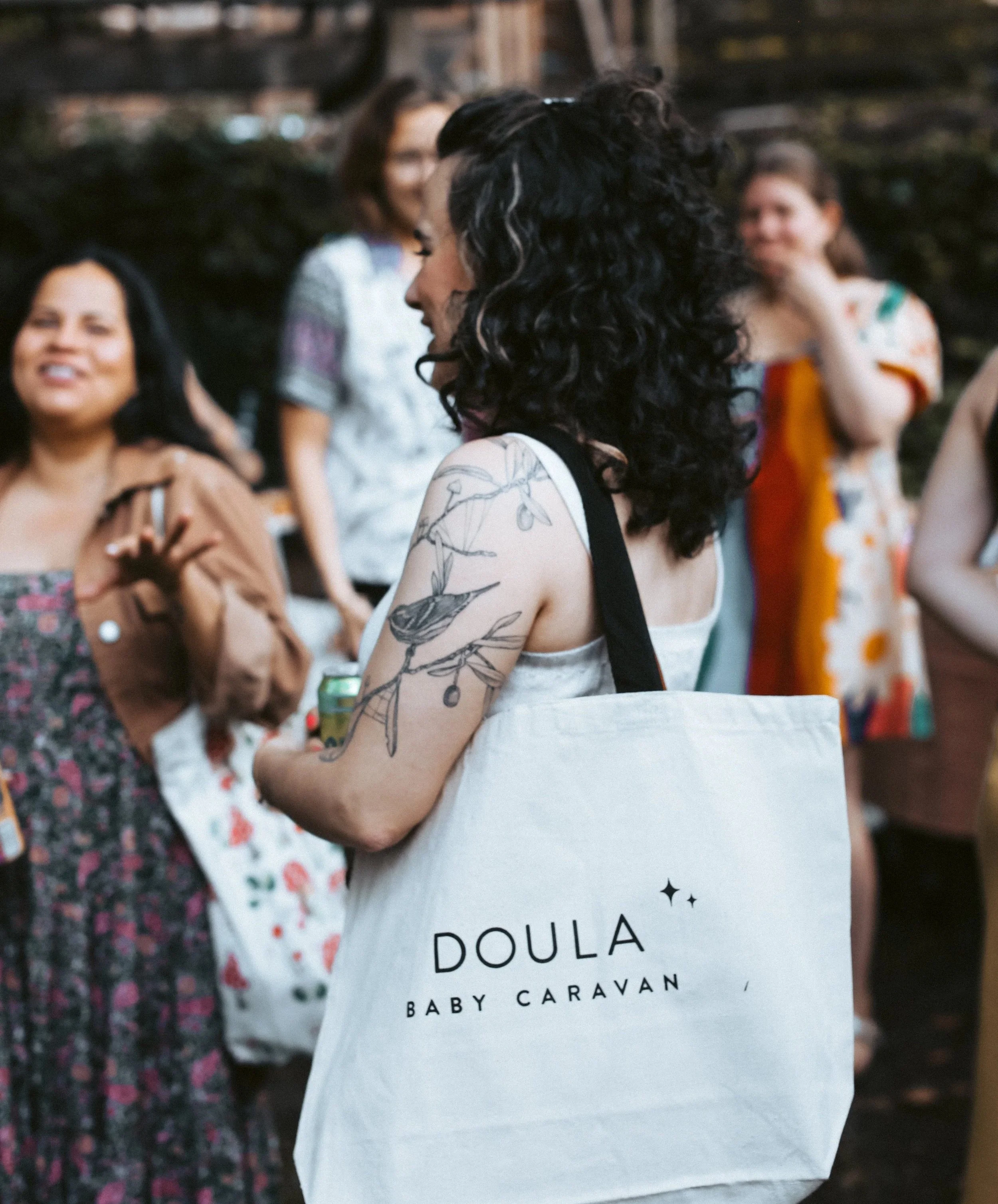 A doula carrying a Baby Caravan branded tote bag at a community gathering, representing Baby Caravan's collective of independent birth and postpartum doulas who run their own businesses in NYC