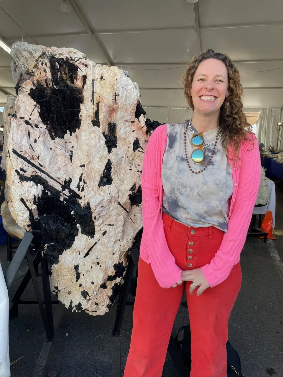 A woman with curly hair and a big smile, wearing a gray tie-dye shirt, bright pink cardigan, and red high-waisted pants, stands next to a large, abstract black and white rock art piece inside a tent.