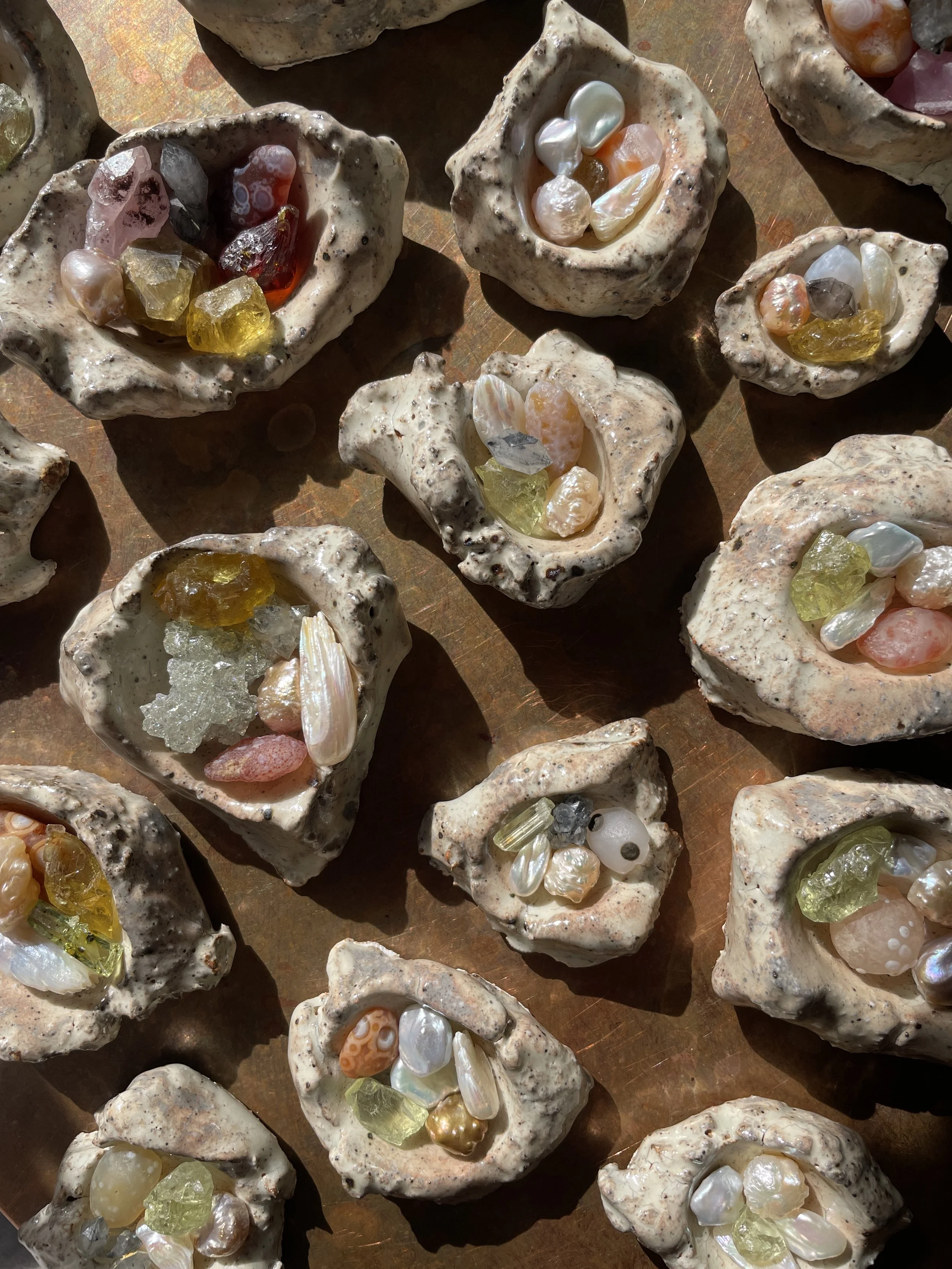 Multiple small handmade pottery bowls filled with various colorful stones, shells, and crystals, arranged on a wooden surface, bathed in sunlight.