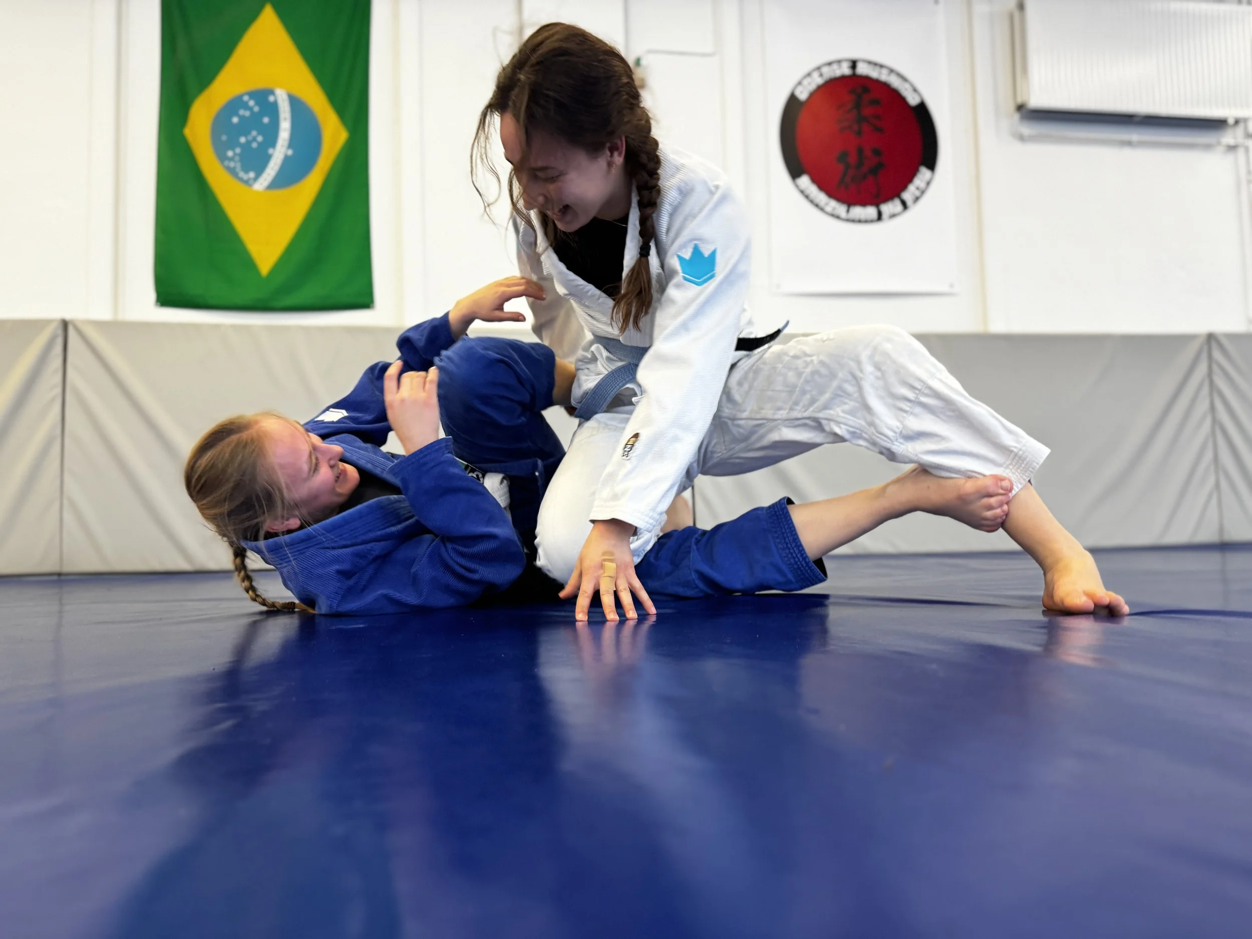 To kvinder træner judo på gulvet, den ene i en sort judodragt, den anden i en hvid. Den med den hvide judodragt er i en position, der forsøger at overvinde den i den sorte judodragt. Baggrund med brasiliansk flag og judo klubskilte.