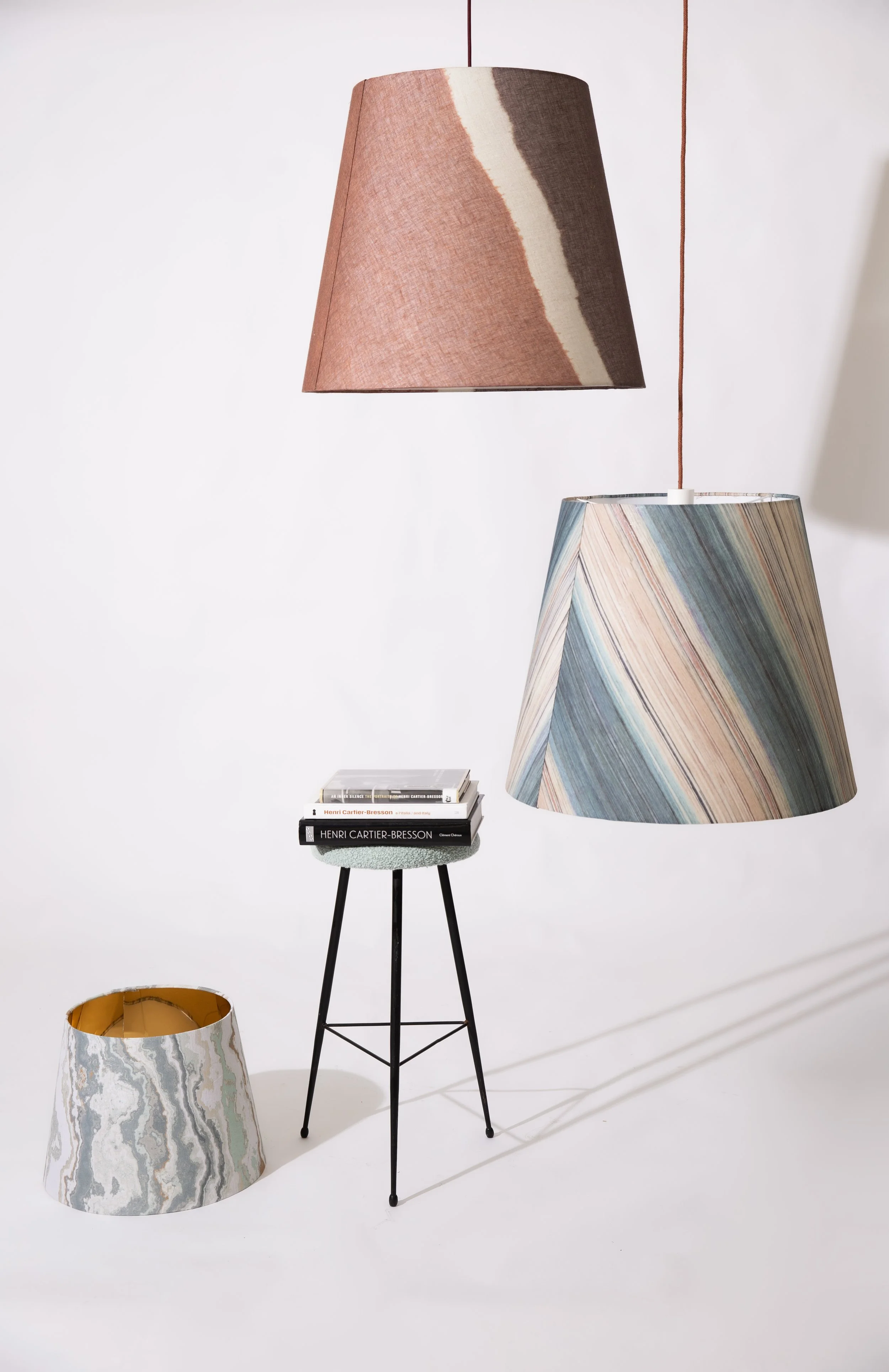 Lampade pendenti con paralumi colorati, una più grande con colori a strisce e una più piccola con tonalità di rosa e marrone, sopra una sedia nera con libri.