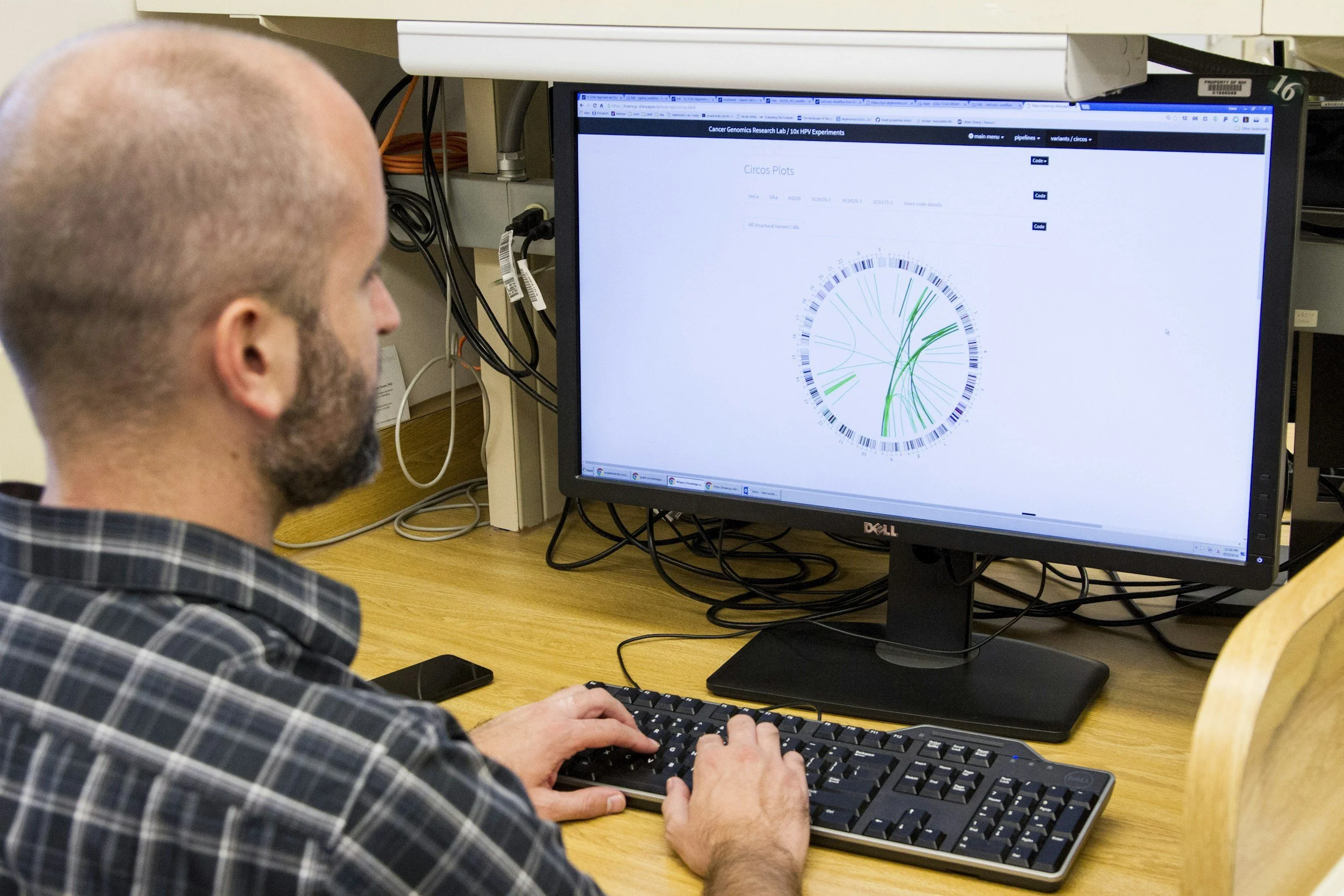 A man with a beard and wearing a plaid shirt working on a computer with a DNA circle plot displayed on the monitor.