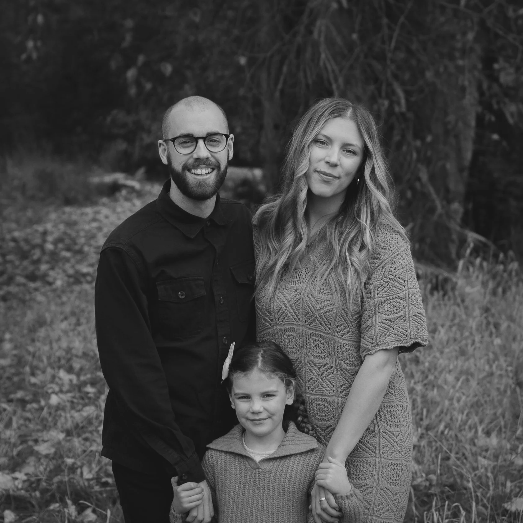 A family of three posing outdoors by the water. The woman in the middle is holding a young girl, and the man is standing beside them. They are smiling and standing near a wooden fence with the ocean and sky in the background.