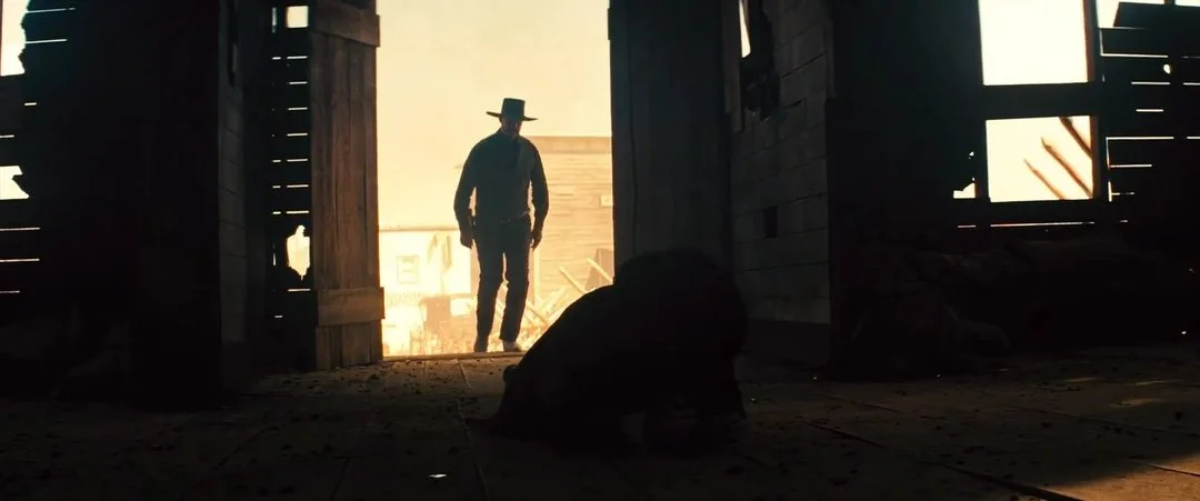 Silhouetted man wearing a wide-brimmed hat walks through a barn doorway toward a figure lying on the floor, with bright sunlight behind him and wooden walls framing the opening.