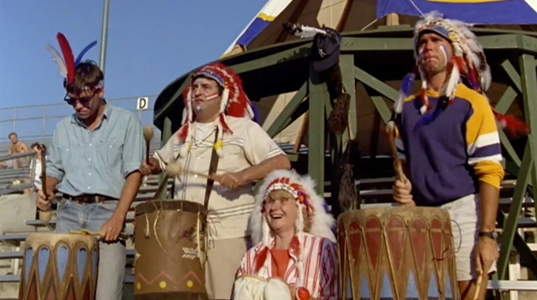 Three white men and a white woman in the bleachers of a baseball game dressed as native Americans and banging on drums with mallets.