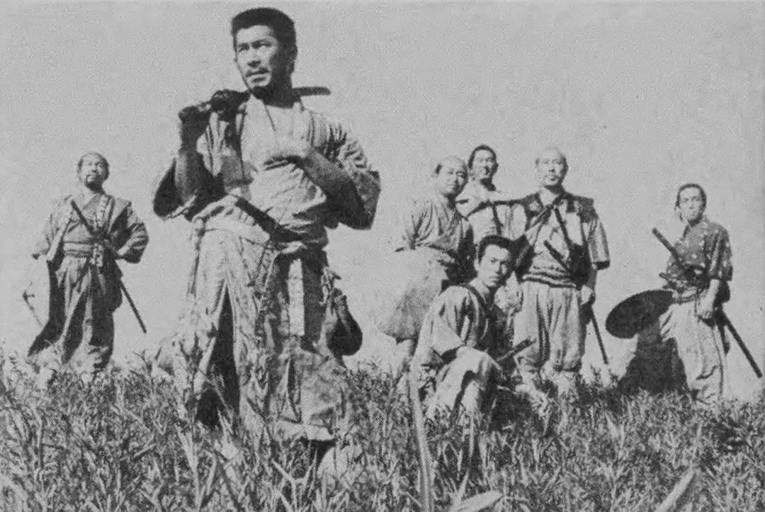 Seven men dressed as samurais stand on a hill of tall grass and look into the distance. black and white.