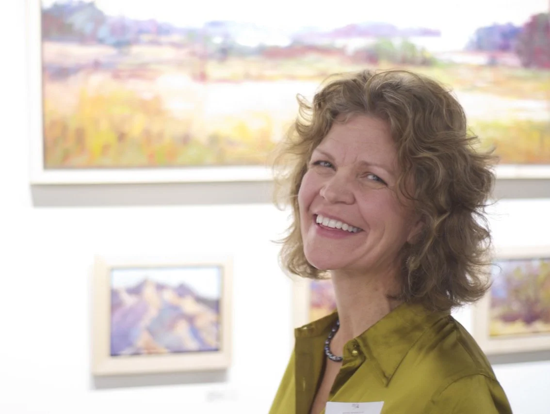 Woman with curly hair smiling in an art gallery with colorful landscape paintings in the background.