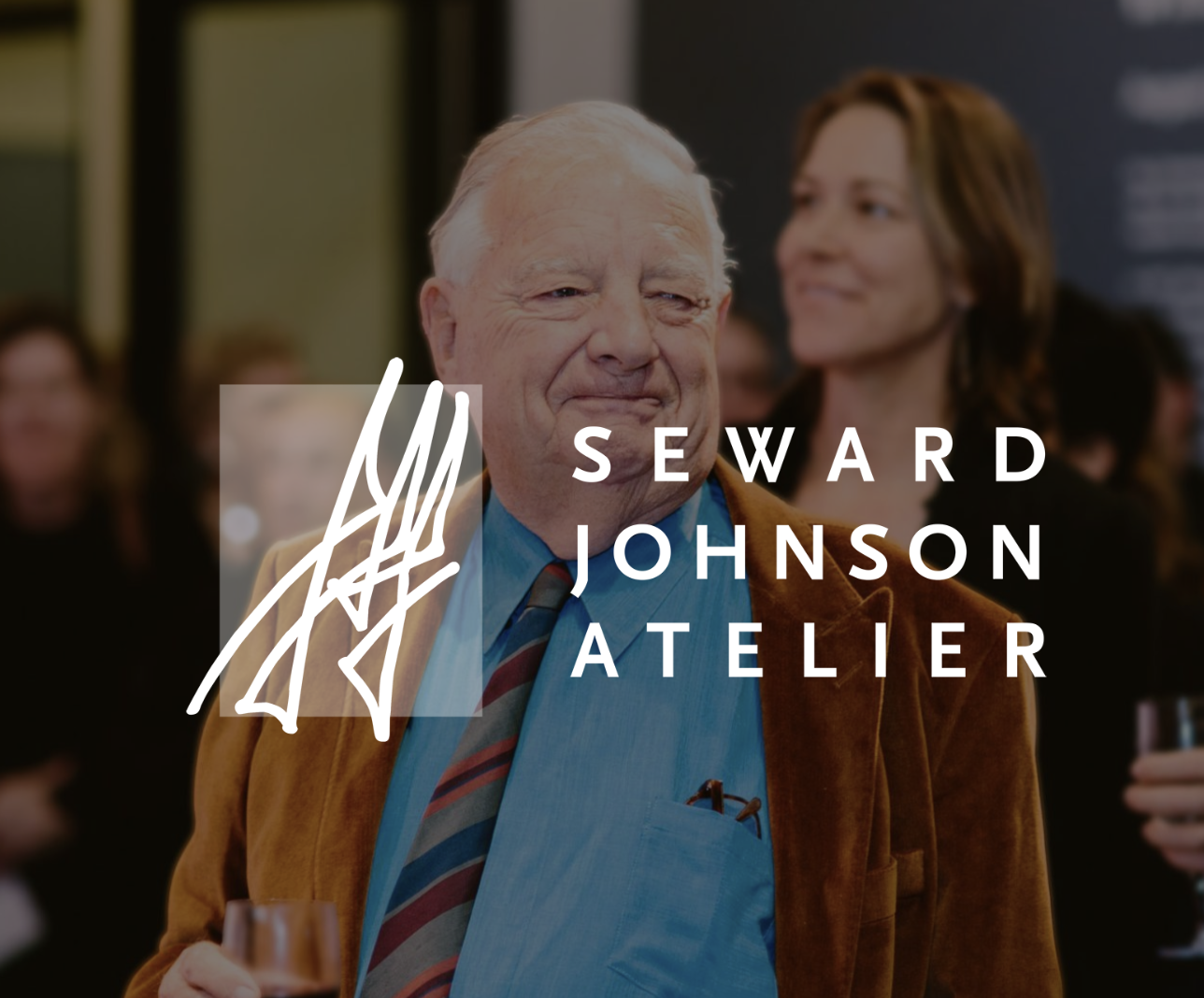 Older man in brown jacket and blue shirt at social gathering with woman in background, logo overlay reading 'Seward Johnson Atelier'.