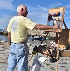 An elderly man painting outdoors on a waterfront with a cityscape and blue sky in the background.