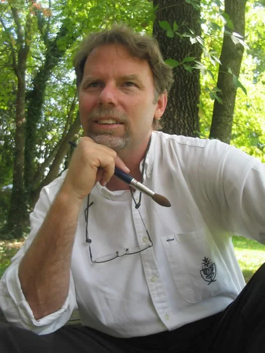 Man with glasses hanging from his neck, holding a paintbrush, sitting outdoors in a wooded area, looking at the camera.