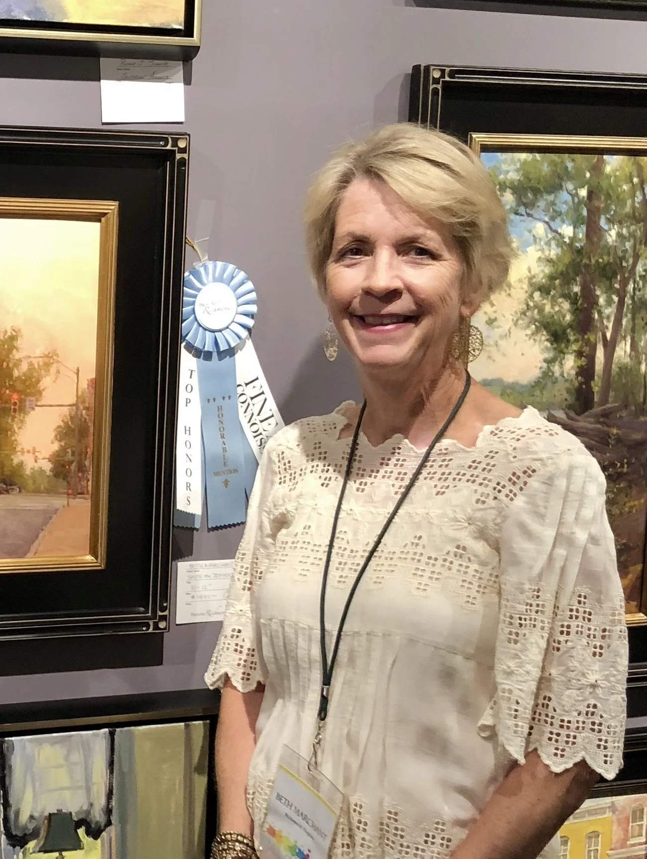 An older woman with short blonde hair smiling at a painting exhibition, wearing a cream-colored embroidered blouse and a black lanyard with her name tag, standing in front of framed landscape paintings and a blue ribbon award.