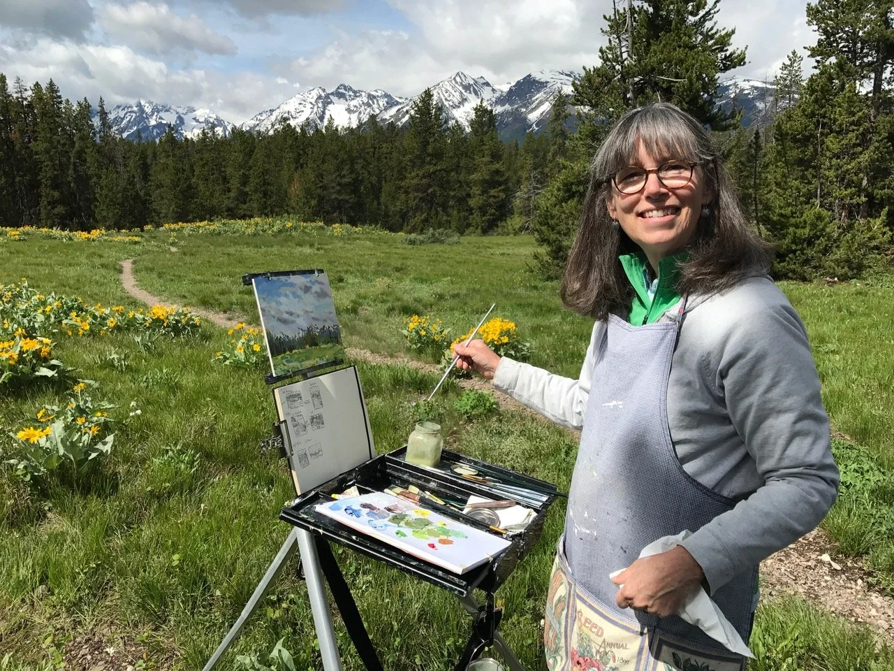 A woman painting outdoors in a green field with yellow flowers, surrounded by trees and snow-capped mountains in the background.