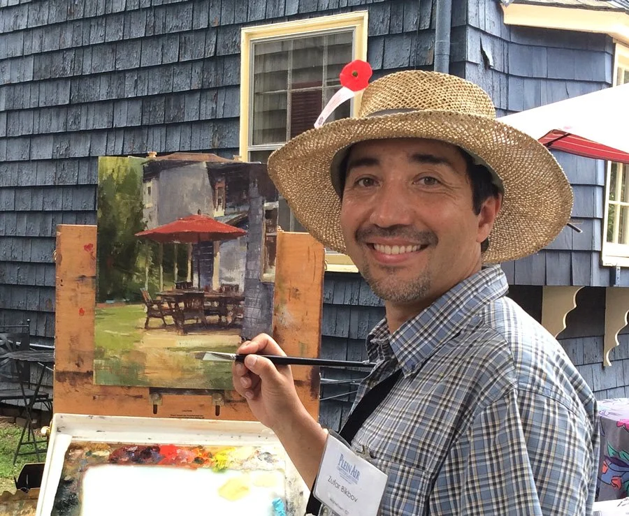 A man wearing a straw hat with a red flower clip, smiling and painting an outdoor scene on a canvas, in front of a blue house with white-trimmed windows.