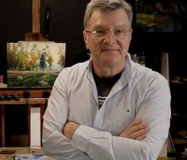 A man with glasses and a white shirt standing with his arms crossed in an art studio, with a landscape painting in the background.