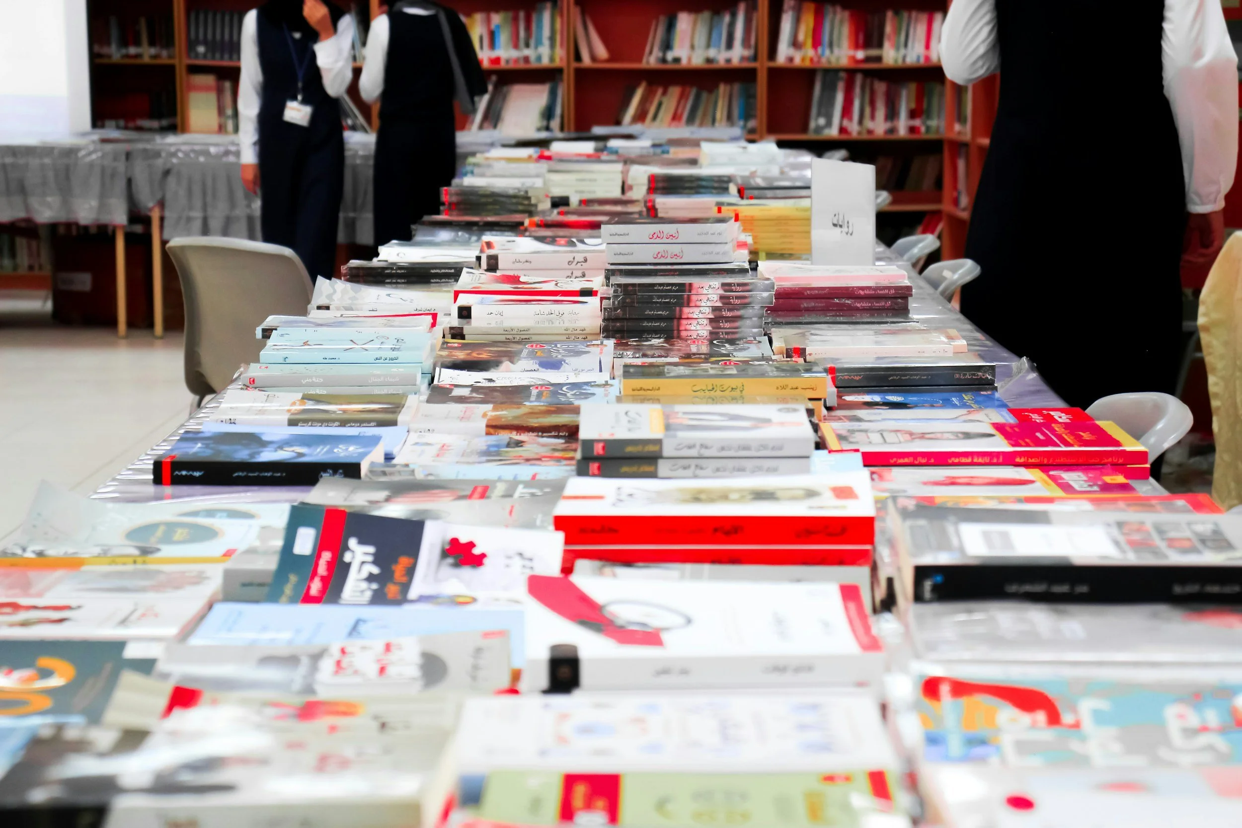 Table filled with stacks of books at a book fair or library.

Background shows bookshelves filled with more books and people browsing or selling.