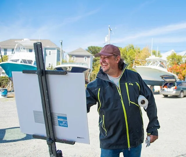 A man standing outdoors near boats, painting on an easel, wearing a dark jacket and cap, smiling.