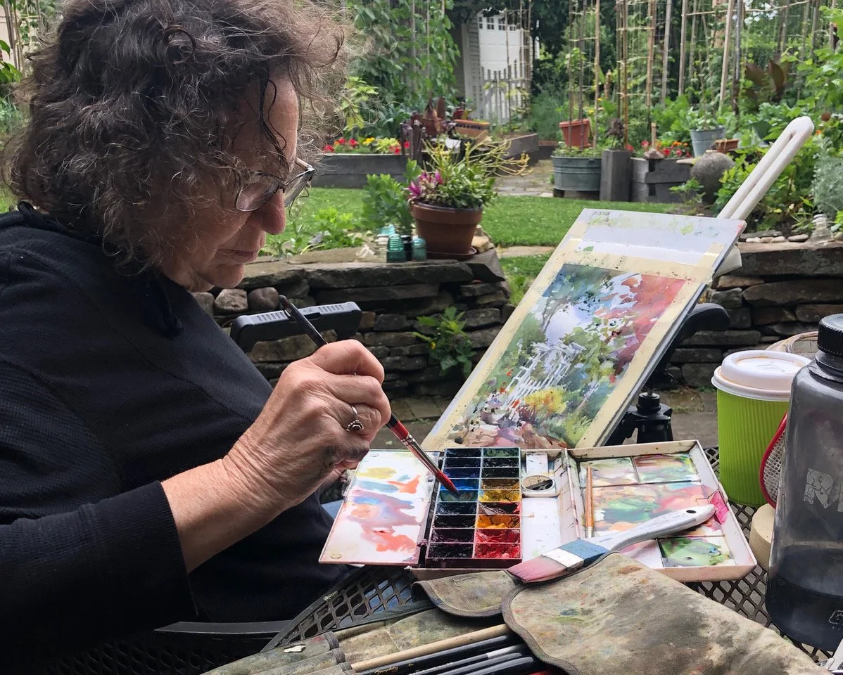 A woman painting watercolor outdoors in a garden. She is sitting at a table with an open watercolor set and a palette, working on a landscape painting of trees and a pathway.