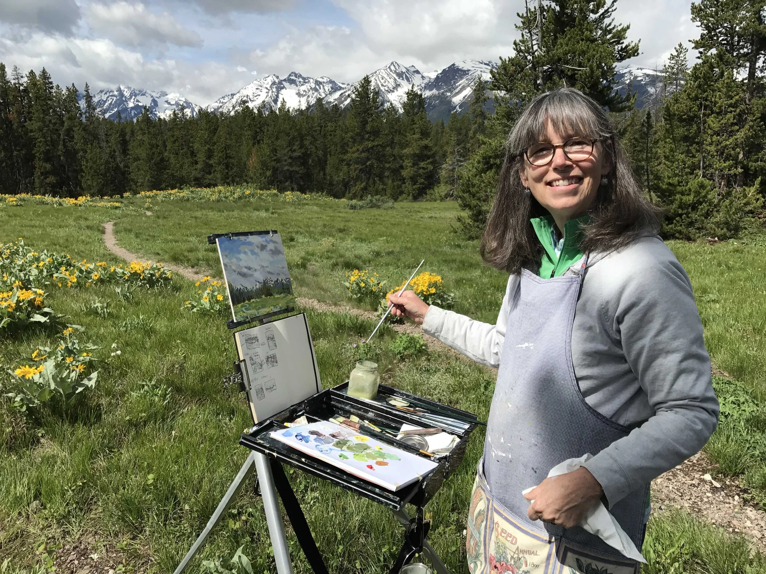 A woman outdoors in a mountainous area painting on a canvas with a sky and trees reflected on an open palette. She is smiling, wearing glasses, a gray apron, and a green jacket, with snow-capped mountains, forests, and yellow flowers in the background.