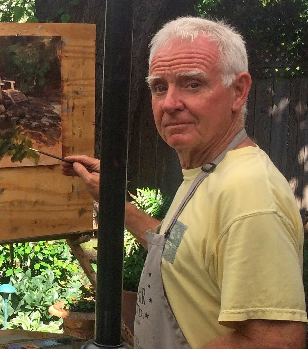 An elderly man stands next to an outdoor painted scene, holding a paintbrush. He is wearing a yellow T-shirt, and a gray apron is hanging around his neck. The background features a wooden fence and potted plants.