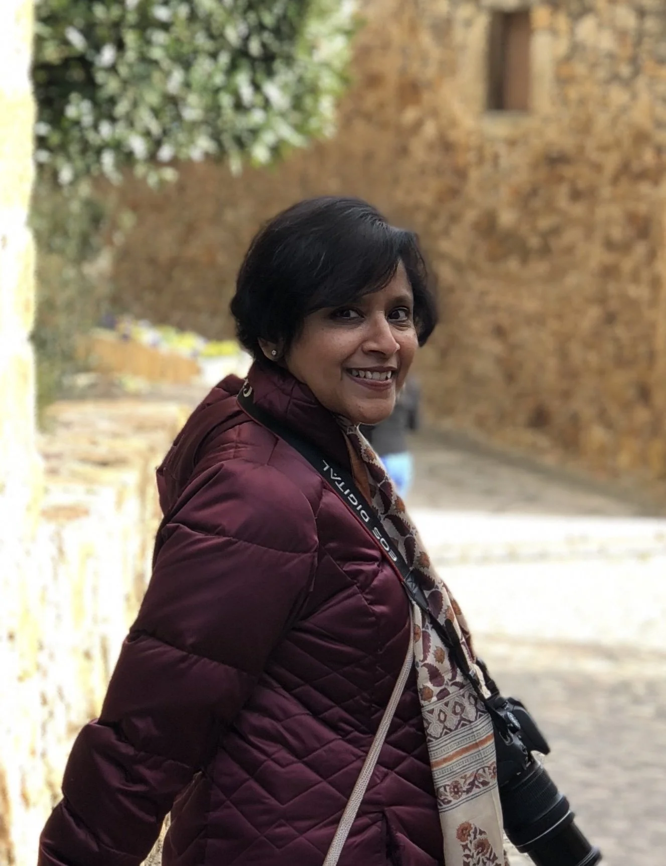 A woman with short black hair smiling outdoors, wearing a maroon quilted jacket, a patterned scarf, and carrying a camera.