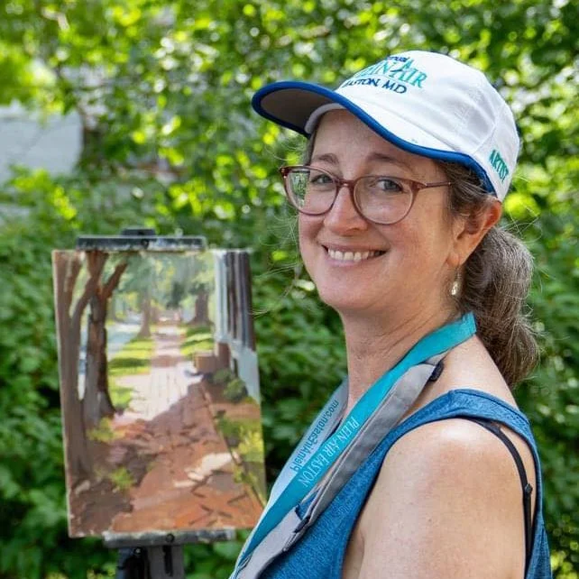 A smiling woman wearing glasses, a white and blue cap, and a blue tank top, standing outdoors in front of an easel displaying a landscape painting of a pathway through trees with green foliage.