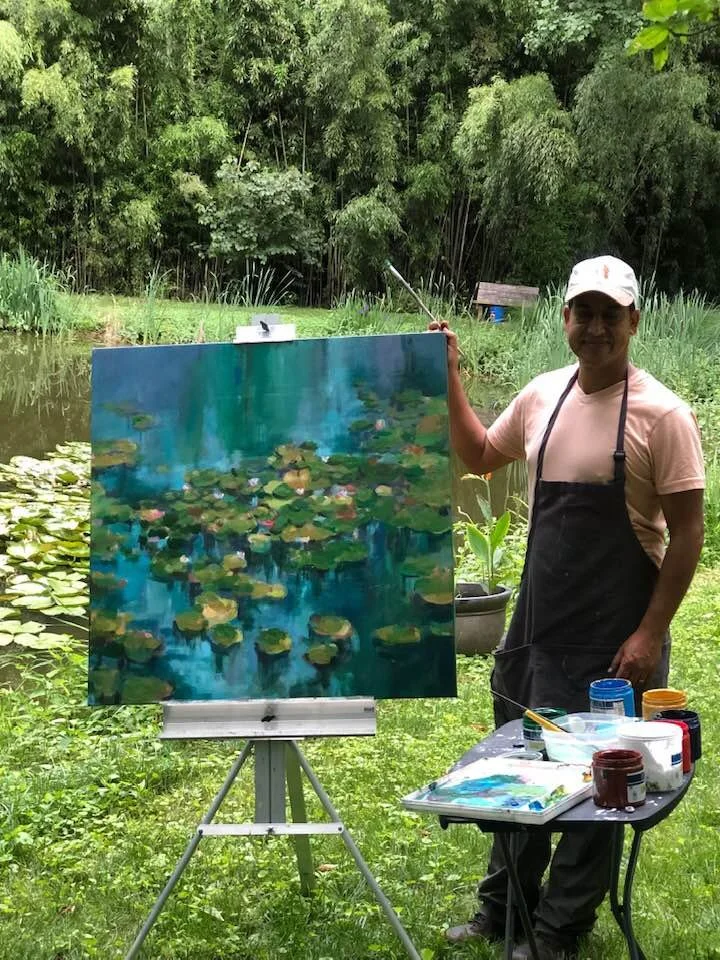 A man standing outdoors near a pond with green lily pads, painting on a canvas set on an easel. He is holding a paintbrush in one hand and is smiling. The scene is lush with trees and grass.