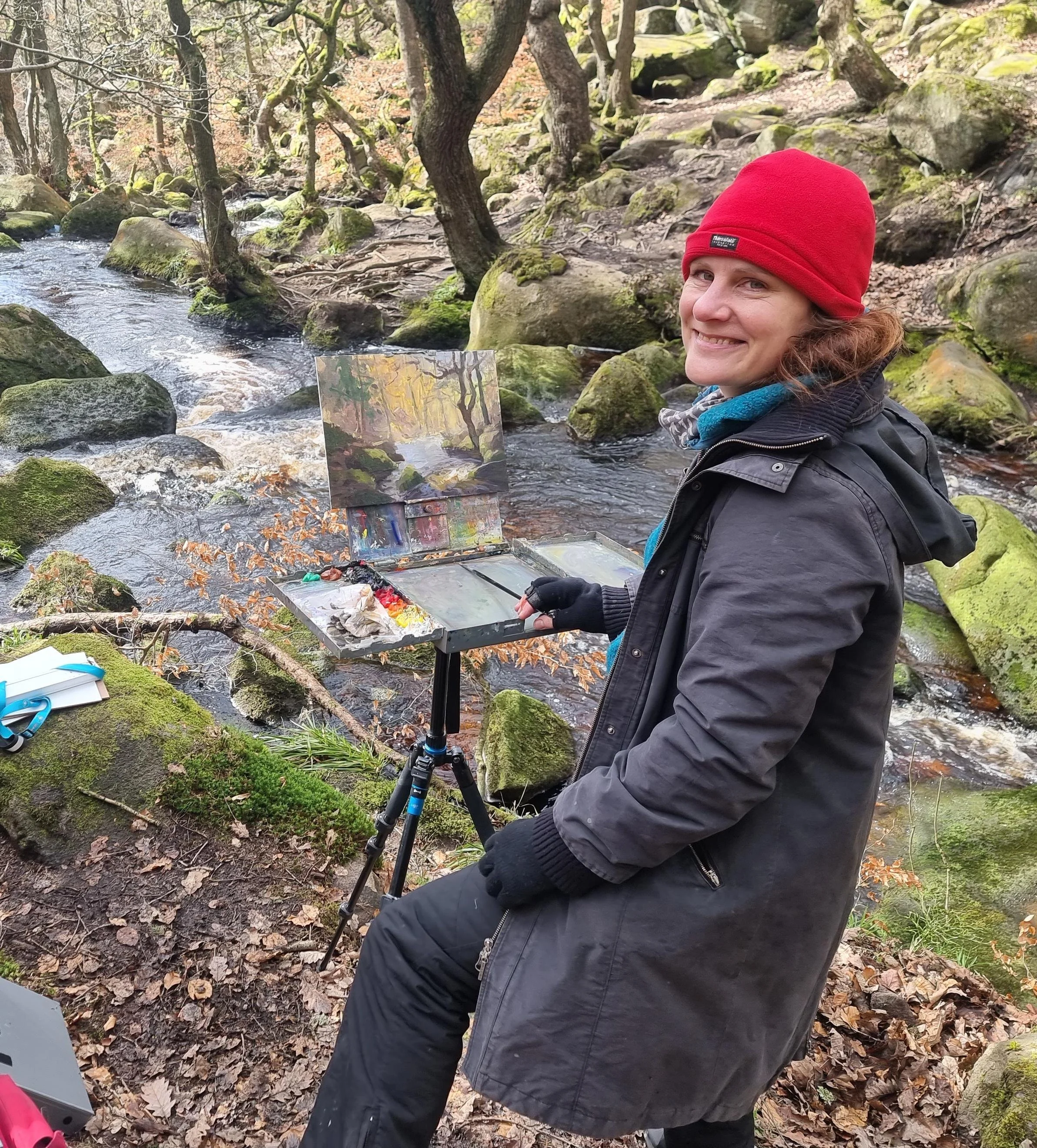 A woman smiling, with an easel and painting supplies in front of her.