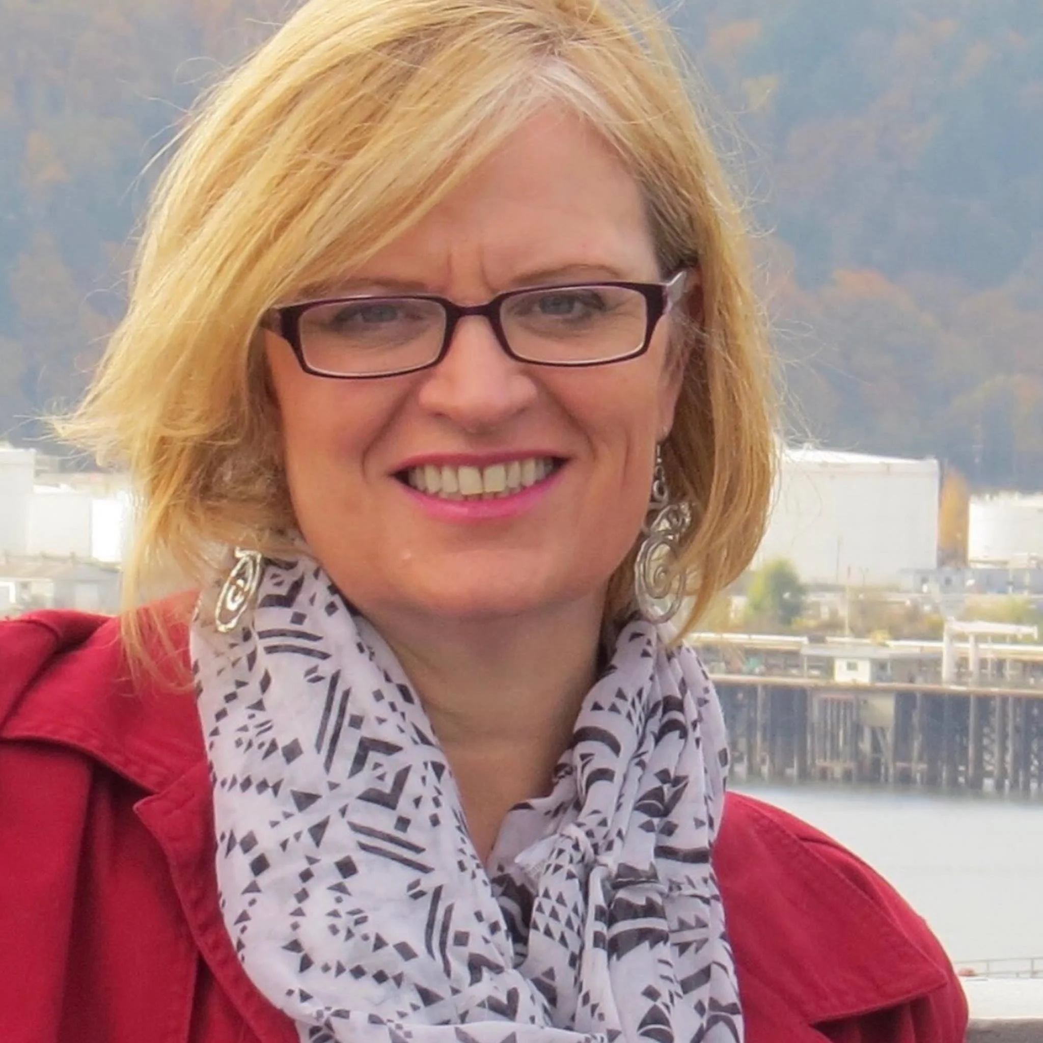 A woman with blonde hair, glasses, and earrings, smiling outdoors near a body of water with a cityscape and hills in the background.