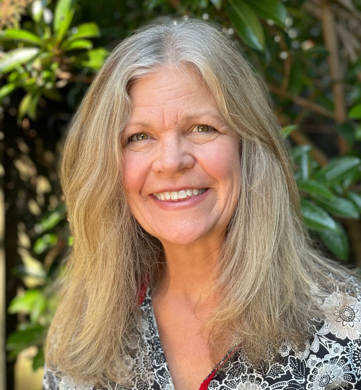 Close-up of a smiling woman with long gray hair, wearing a black and white floral top, outdoors with green foliage in the background.