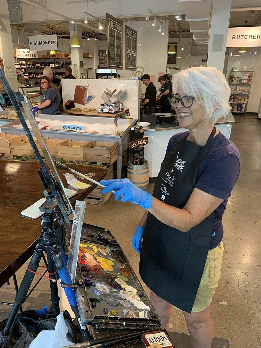 A woman with short gray hair, glasses, and a black apron is painting on a canvas set on an easel at an indoor market or store. She is smiling and wearing blue gloves, with a palette of mixed paints in front of her. In the background, there are other people shopping or working at various counters and shelves, including a butcher section and wine section.