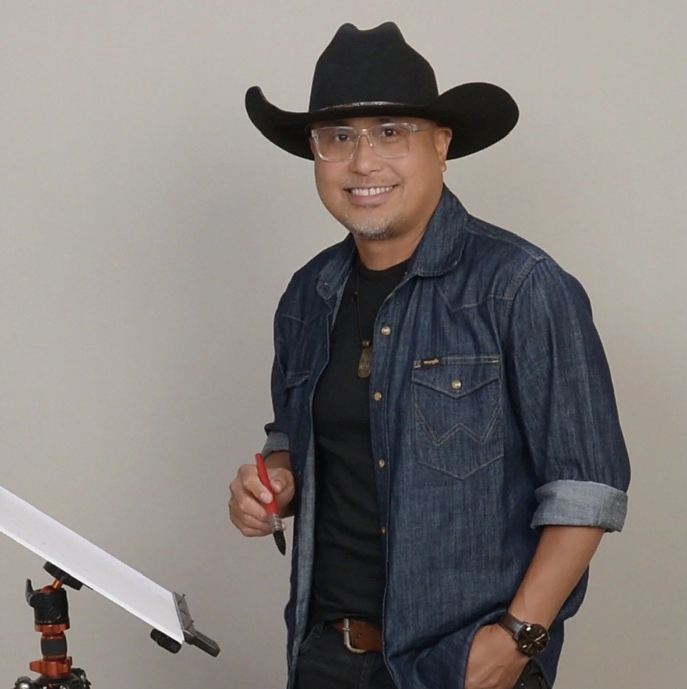A man wearing a cowboy hat and dark clothing stands outdoors on a sunny day, painting a desert landscape on a small easel. Behind him are desert hills, a few structures, and sparse vegetation, with a shed to the right.