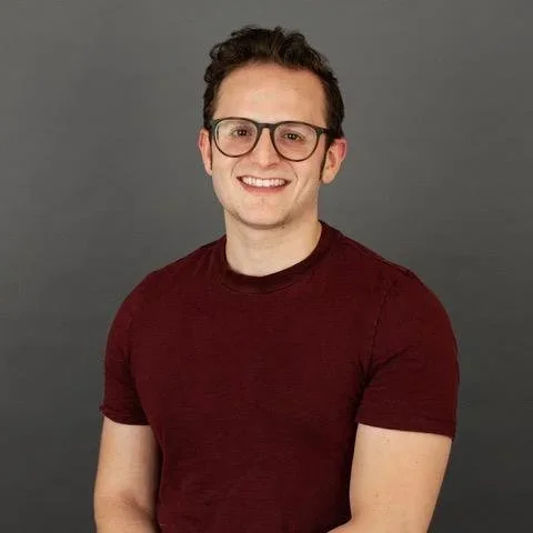 Jeff Lichtenstein with glasses smiling, wearing a maroon t-shirt, with short dark curly hair, against a gray background.