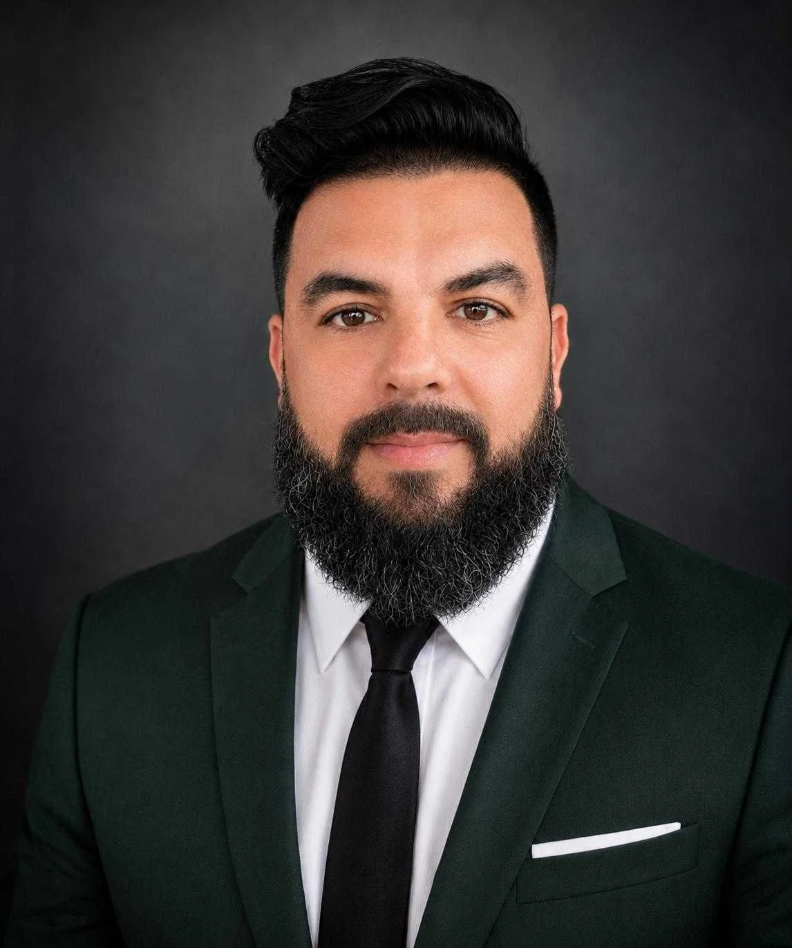 Portrait of a man with dark hair and beard wearing a dark green suit, white shirt, and black tie, against a dark background.
