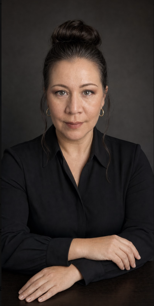Portrait of a woman with dark hair in a bun, wearing gold hoop earrings and a black shirt, sitting with crossed arms against a dark background.