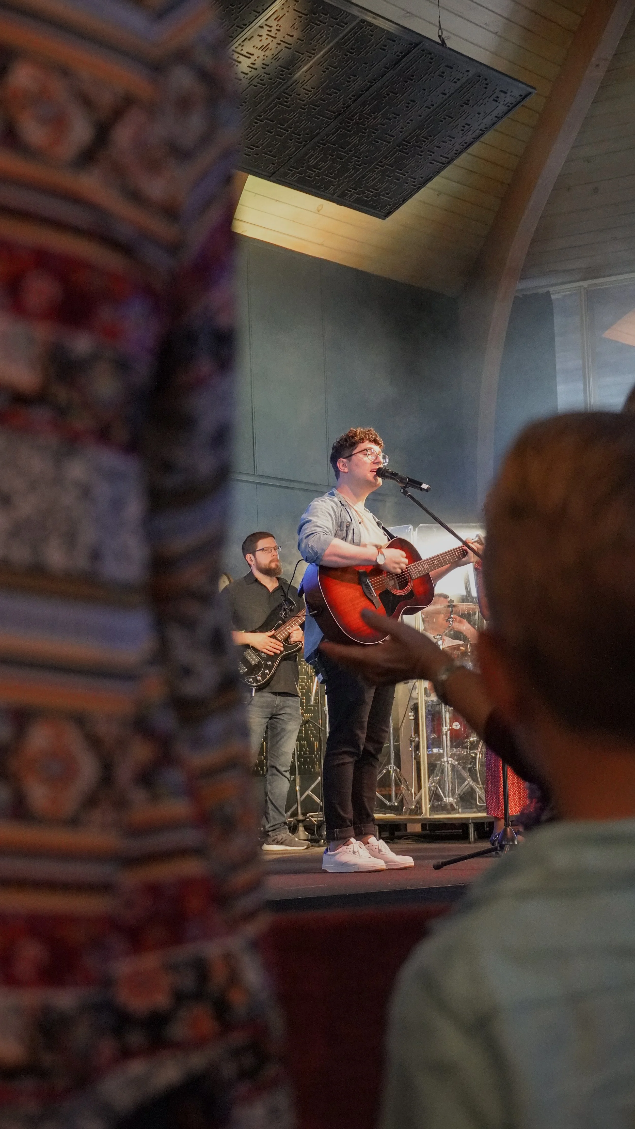 Musician playing guitar and singing on stage, with band members in the background, during a live performance at an indoor venue.