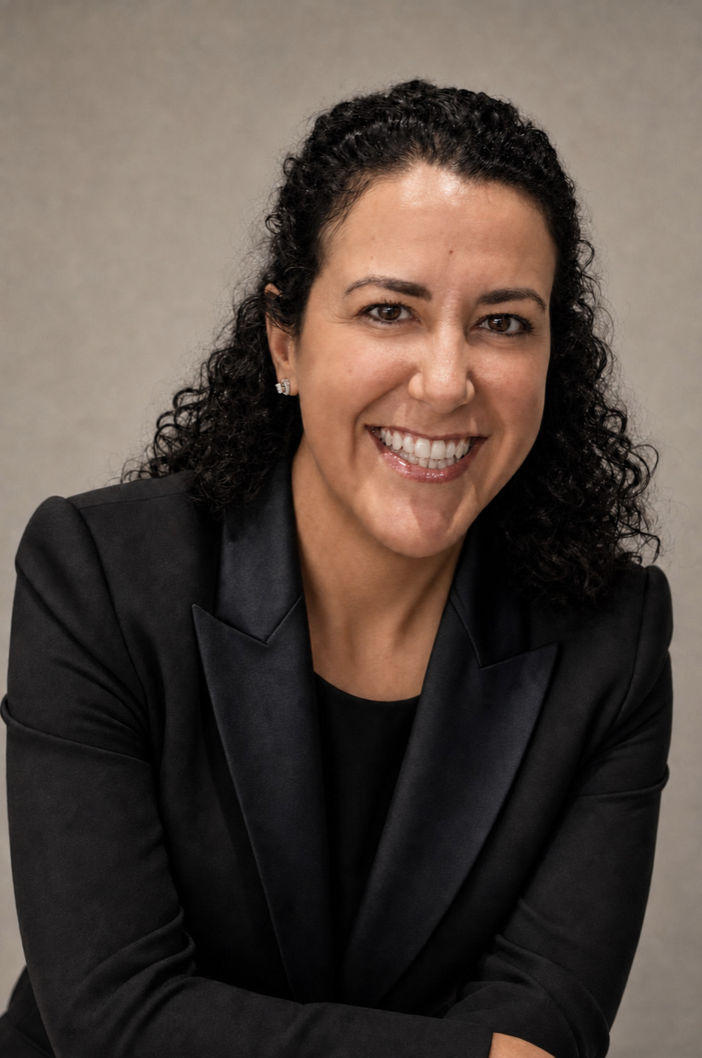 A woman with curly dark hair smiling and wearing a black blazer and top, sitting against a neutral background.