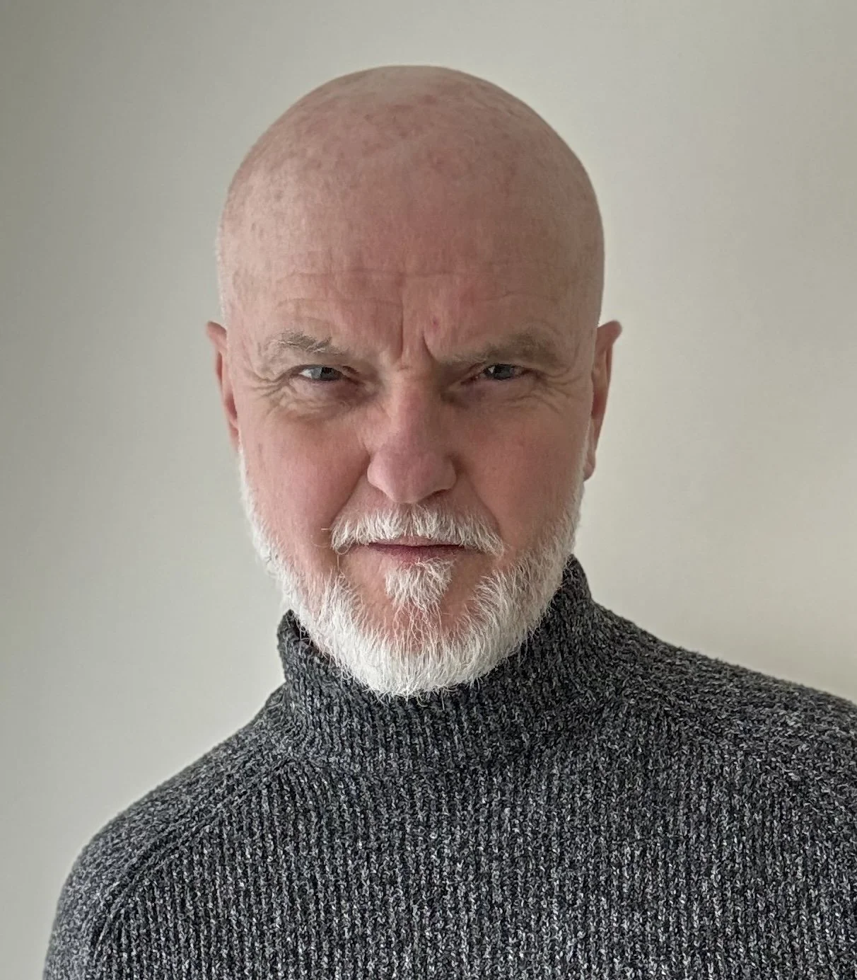A close-up portrait of teapotmonk o with a bald head, white beard, and wearing a dark gray knit turtleneck sweater, standing against a plain light-colored background.