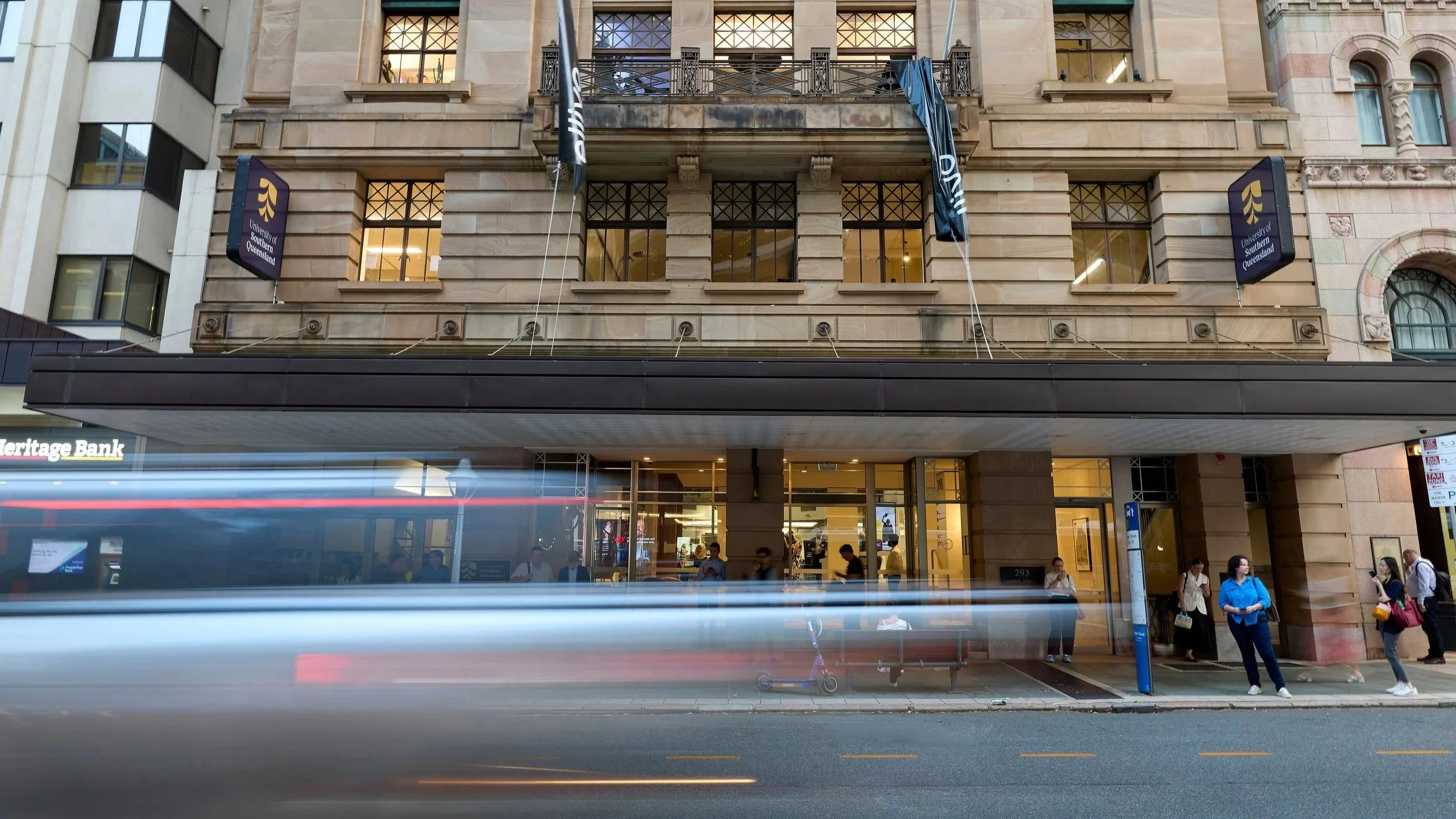 People waiting outside a building of the University of Southern Queensland, with a blurred bus or train passing by on the street.